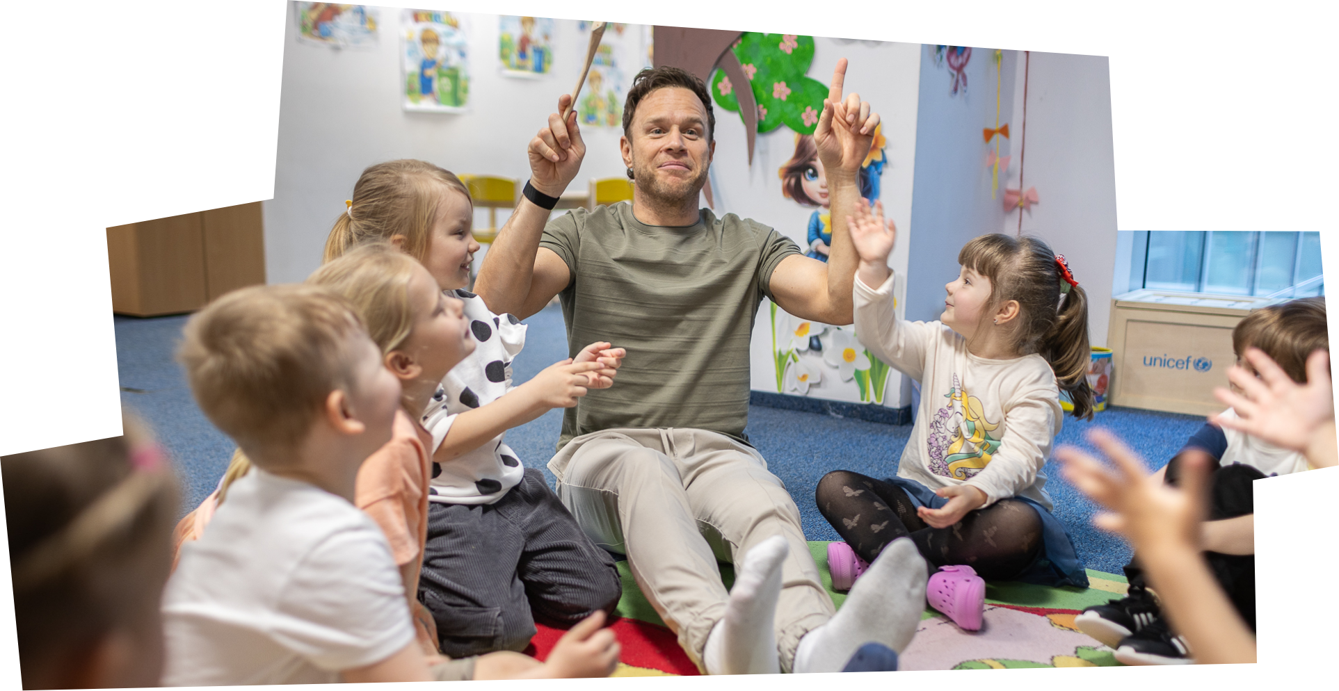 Singer Olly Murs sits on the floor of a children's play centre in Bucharest. He is surrounded by young Ukrainian children who are sitting with their legs crossed in a circle.